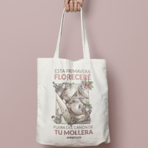 En la imagen se observa una foto de producto, el producto es una tote bag, es decir, una bolsa de tela color crudo (blanco natural) con una ilustración dibujada donde se ve un cuerpo encuadrado en un rectángulo con pechos medianos, michelines, celulitis y pelos. Esta dibujo está acompañado por botánica entre flores y hojas, y en el texto pone "Esta primavera floreceré fuera del canon de tu mollera" La ilustración es obra de Eva Cortés Ilustra