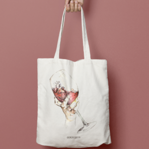 En la imagen se observa una foto de producto, el producto es una tote bag, es decir, una bolsa de tela color crudo (blanco natural) con una copa de vino dibujada con estilo lápiz y acuarelas. Esta copa está sostenida por una mano de forma delicada, y dentro de ella, con el vino, hay un corazón anatómico. La ilustración es obra de Eva Cortés Ilustra