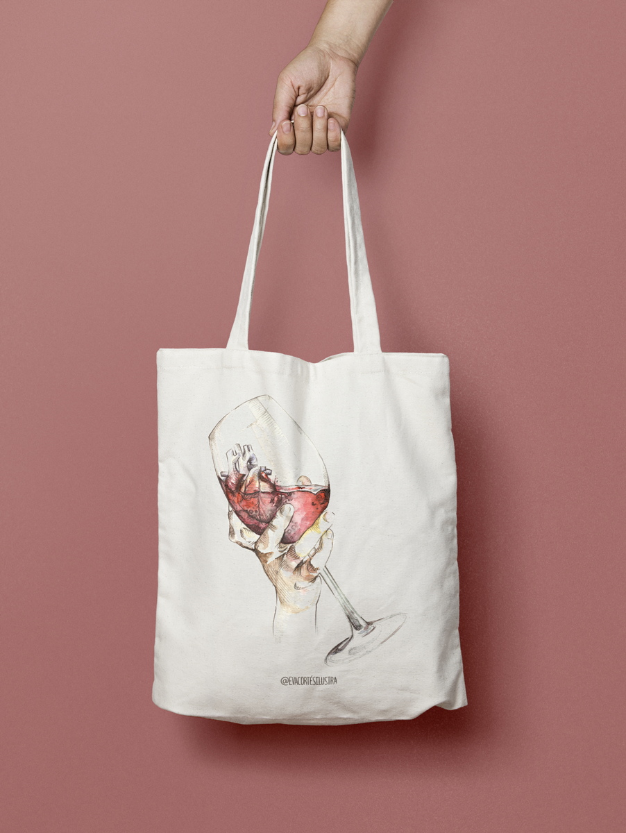 En la imagen se observa una foto de producto, el producto es una tote bag, es decir, una bolsa de tela color crudo (blanco natural) con una copa de vino dibujada con estilo lápiz y acuarelas. Esta copa está sostenida por una mano de forma delicada, y dentro de ella, con el vino, hay un corazón anatómico. La ilustración es obra de Eva Cortés Ilustra