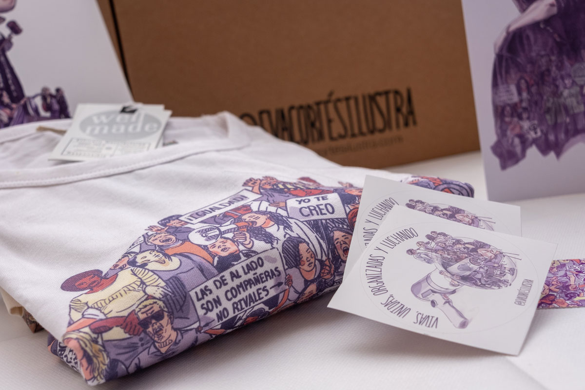 En la imagen se ve al detalle y de cerca el tipo de impresión (digital textil) de la camiseta que forma parte de este pack de productos de temática feminista "8 de marzo". En la imagen se puede observar de cerca la calidad del tejido de la camiseta, la cual cumple con parámetros éticos de producción textil. Al lado se ven dos pegatinas que se incluyen de regalo en el pack de Eva Cortés.