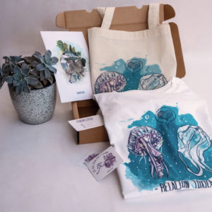 En la imagen se ve un ejemplo del pack de productos de temática feminista "ecologista". En la imagen se puede observar una lamina DIN A5 con una de las ilustraciones que reflexionan sobre nuestro planeta de Eva Cortés, una totebag y una camiseta de la misma línea.