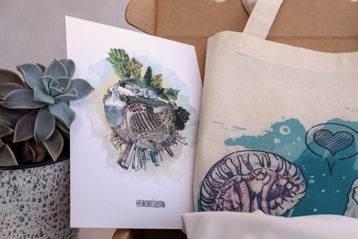 En la imagen se ve al detalle y de cerca el tipo de impresión de la lámina pequeña que forma parte de este pack de productos de temática feminista "ecologista". En la imagen se puede observar una lamina pequeña, apoyada en una planta suculenta, junto a una bolsa tote bag con uno de los conocidos diseños de "relación toxica" de Eva Cortés.