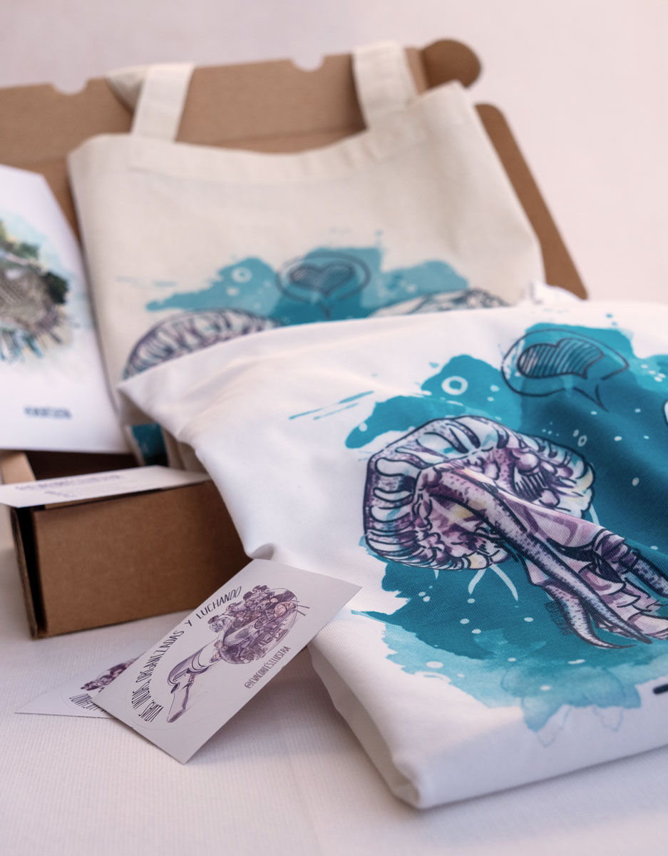 En la imagen se ve un ejemplo del pack de productos de temática feminista "ecologista". En la imagen se puede observar más de cerca el detalle de la impresión digital textil de la camiseta "Relación Tóxica", junto con unas pegatinas , y de fondo una totebag y una lamina DIN A5 de la misma línea.