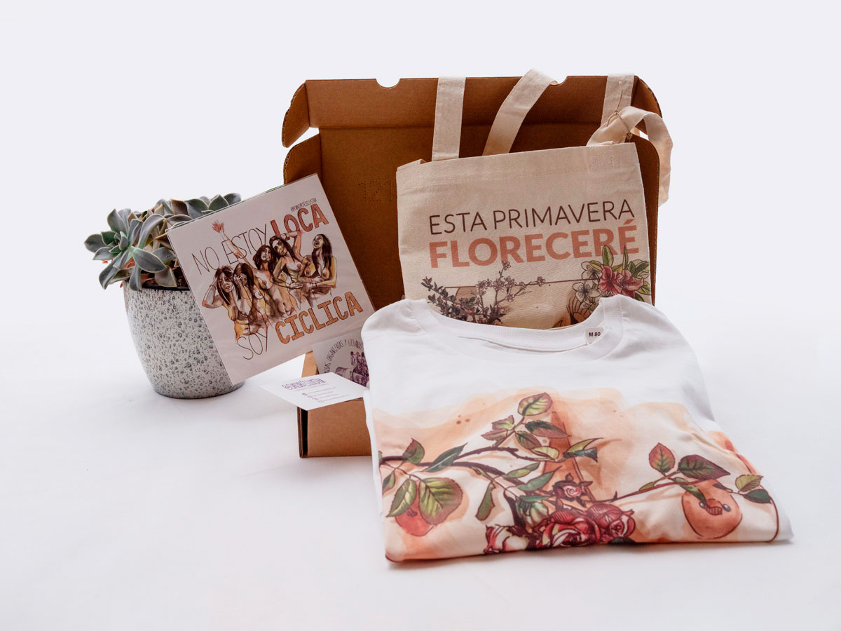 En la imagen se ve un ejemplo del pack de productos de temática feminista "Cuerpos Diversos". En la fotografía se puede observar una lamina pequeña DIN A5 (el pack incluye dos) que se titula "No estoy loca, soy cíclica" , también se ve una camiseta con uno de los conocidos diseños de pechos de Eva Cortés, y una bolsa de tela (tote bag)con la ilustración "Esta primavera floreceré, fuera del canon de tu mollera"